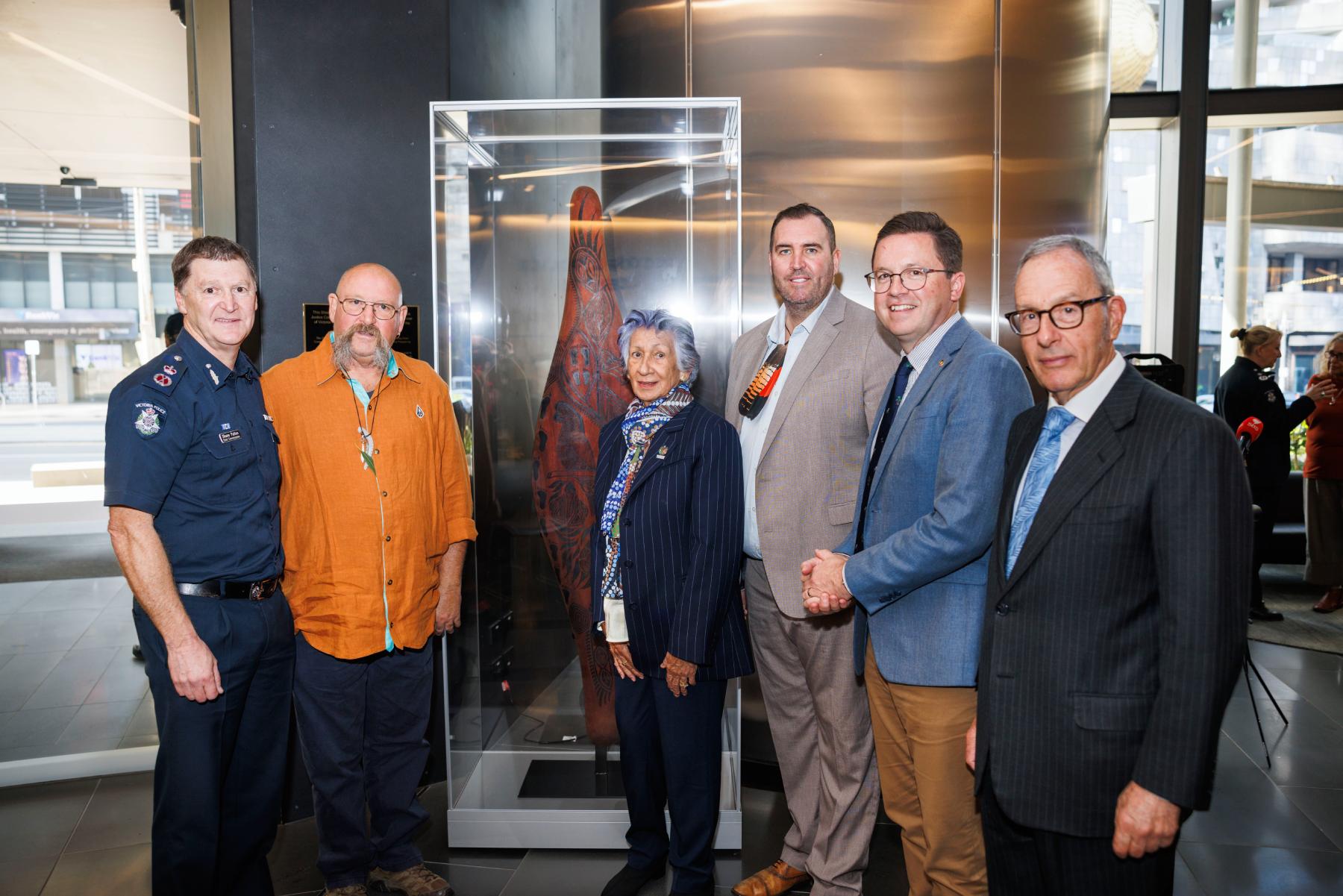 Left to right:  former Chief Commissioner of Police, Shane Patton, Uncle Mick Harding, Yoorrook Justice Commission Chair Professor Eleanor Bourke AM, Yoorrook Justice Commissioner Travis Lovett, Hon Anthony Carbines MP, Minister for Police, Yoorrook Justice Commissioner The Hon. Anthony North KC in the foyer of the Victoria Police Centre. The Shield is made of wood with intricate Aboriginal carvings and is displayed in a glass cabinet central to the image.