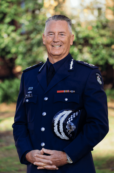 Mike Bush CNZM, Chief Commissioner of Victoria Police, is smiling while standing in a garden and wearing his dark blue police dress uniform. Mike has his hands clasped in front him and his peaked police cap under one arm. 