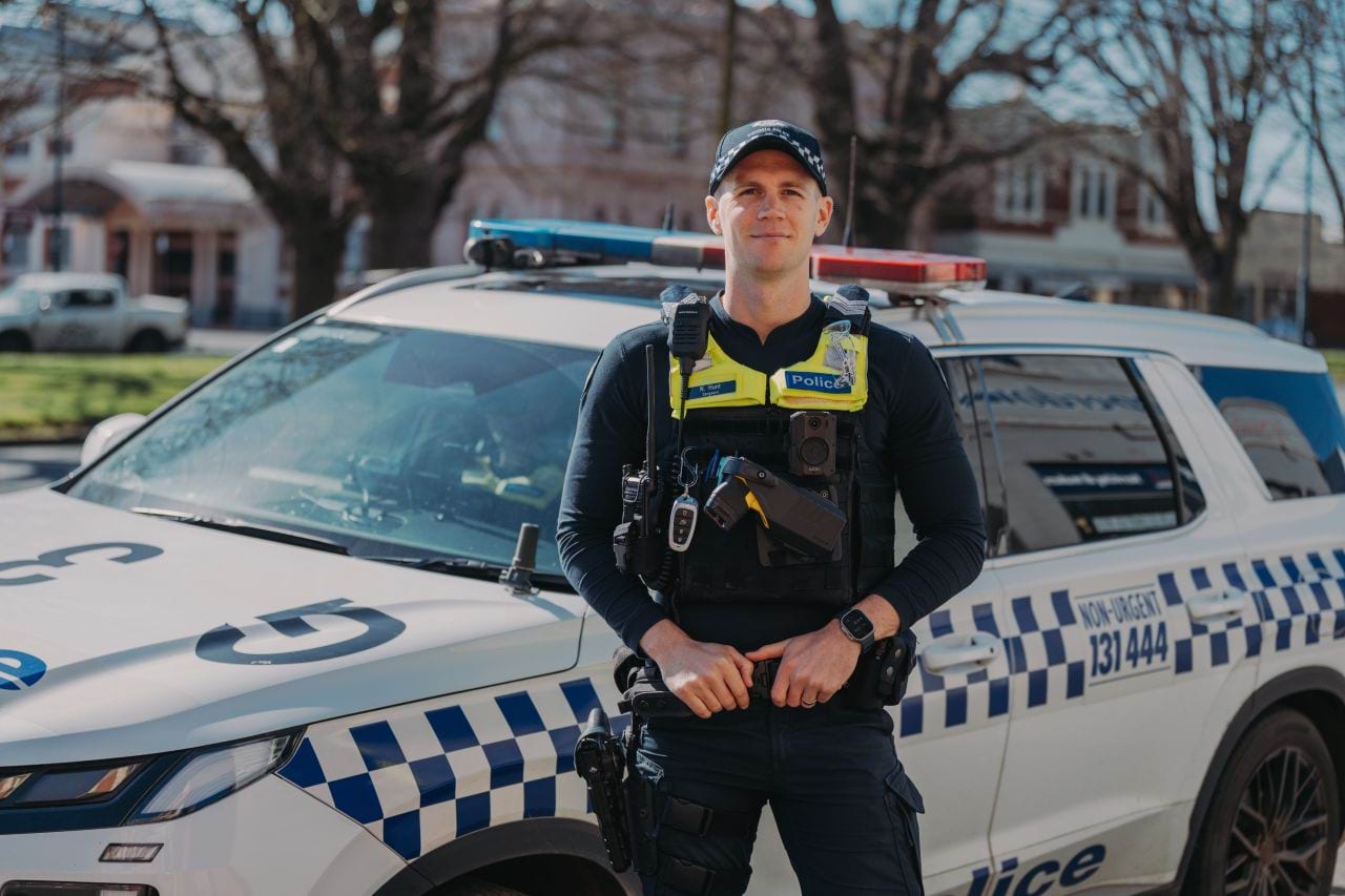 Uniformed police officer standing in the sunshine in front of a police divisional van. He has a slight smile and is looking directly at the camera.  